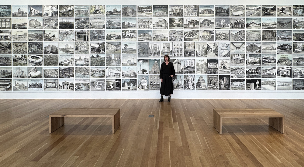 artist Ellen Harvey stanging in a gallery oin front of a wall filled with hundreds of small drawings